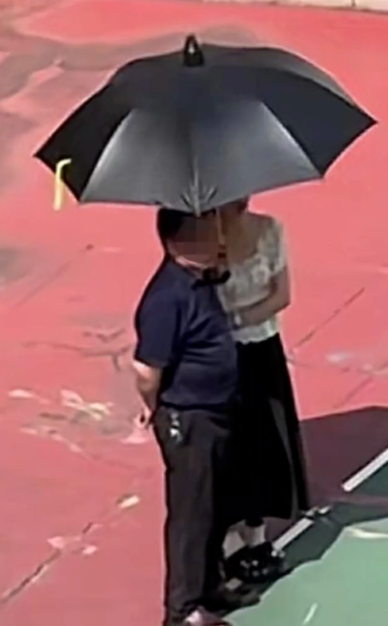 廈門市湖里區世紀學校一群學生於烈日下在操場列隊參加活動,一名女教師在場邊為身旁背手站立的男子撐傘擋陽光。(網上圖片)