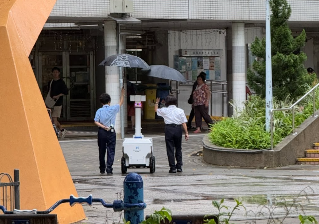 社交平台近日流傳相片，兩名疑似公屋保安於下雨時為一部機械人擔遮，據指該巡邏機械人為本地一間創科平臺企業研發，本身可「防暴雨」。(Threads)